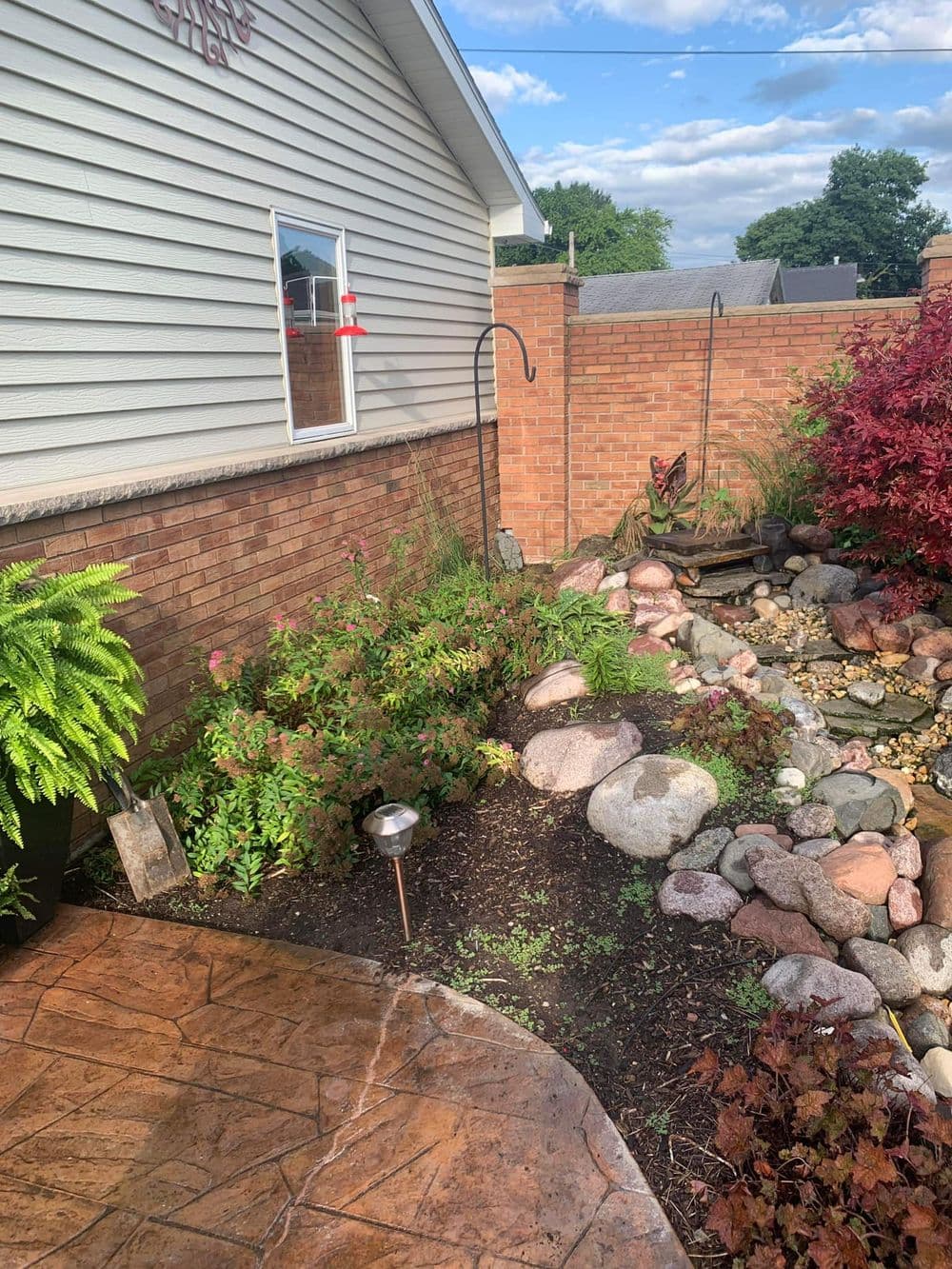 Lush garden with a stone pathway, pond, and decorative plants beside a house.